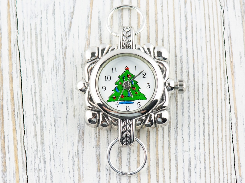 May include: Silver charm watch with a Christmas tree design on the face. The watch has a square face with a silver metal frame and a silver chain.