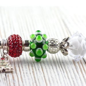May include: A festive bracelet featuring a red, sparkling bead, a green and white glass bead, and a clear, faceted bead. A silver charm in the shape of a Christmas tree hangs from the bracelet, perfect for the holiday season.