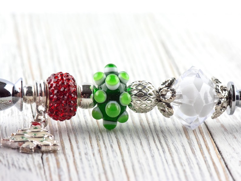 May include: A festive bracelet featuring a red, sparkling bead, a green and white glass bead, and a clear, faceted bead. A silver charm in the shape of a Christmas tree hangs from the bracelet, perfect for the holiday season.