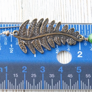 K&ouml;nnte beinhalten: Ein Paar Ohrringe mit einem Farnblatt-Design und einer gr&uuml;nen Edelsteinperle. Die Ohrringe sind aus Metall und etwa 5 cm lang.