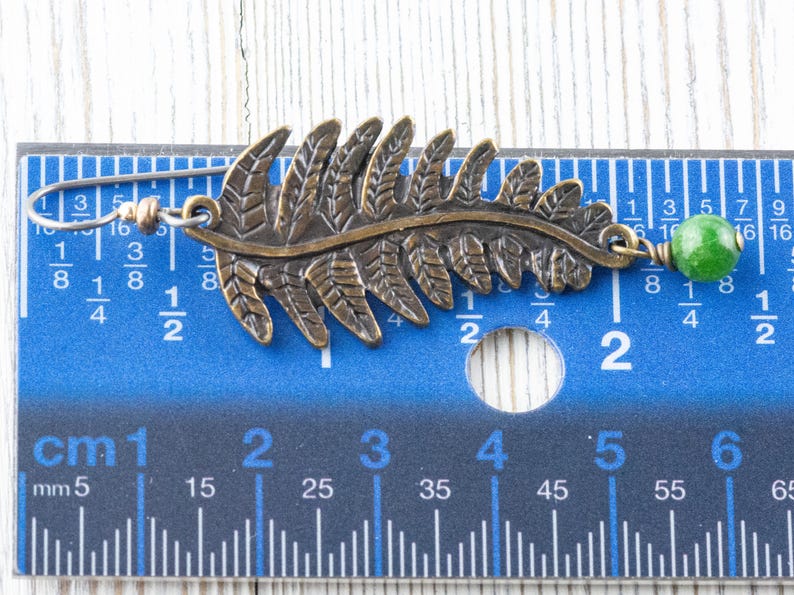K&ouml;nnte beinhalten: Ein Paar Ohrringe mit einem Farnblatt-Design und einer gr&uuml;nen Edelsteinperle. Die Ohrringe sind aus Metall und etwa 5 cm lang.