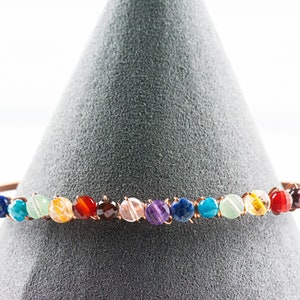 K&ouml;nnte beinhalten: Ein Kupferdraht-Armband mit runden, bunten Edelsteinen. Das Armband zeigt einen Regenbogen aus Farben, darunter Lila, Blau, Gr&uuml;n, Gelb, Orange und Rot.