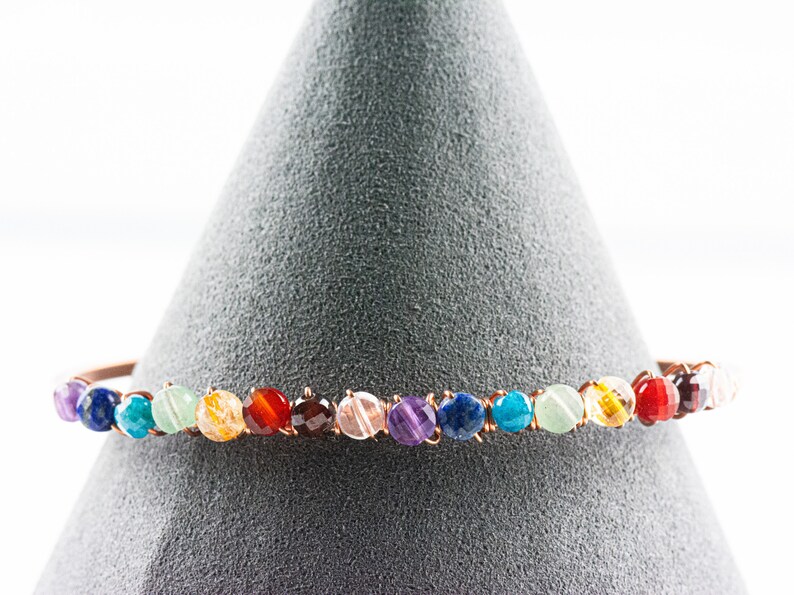 K&ouml;nnte beinhalten: Ein Kupferdraht-Armband mit runden, bunten Edelsteinen. Das Armband zeigt einen Regenbogen aus Farben, darunter Lila, Blau, Gr&uuml;n, Gelb, Orange und Rot.