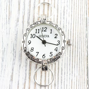 Pode incluir: Rel&oacute;gio de bolso prateado com mostrador branco e algarismos pretos. O rel&oacute;gio tem uma moldura decorativa e dois an&eacute;is para fixa&ccedil;&atilde;o. A palavra "GENEVA" est&aacute; impressa no mostrador. Um acess&oacute;rio decorativo.