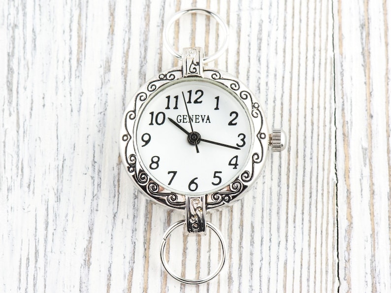 Pode incluir: Rel&oacute;gio de bolso prateado com mostrador branco e algarismos pretos. O rel&oacute;gio tem uma moldura decorativa e dois an&eacute;is para fixa&ccedil;&atilde;o. A palavra "GENEVA" est&aacute; impressa no mostrador. Um acess&oacute;rio decorativo.