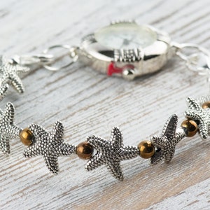 K&ouml;nnte beinhalten: Eine silberne Uhr mit wei&szlig;em Zifferblatt und einem silbernen Armband, das mit Seestern-Anh&auml;ngern und bronzenen Perlen verziert ist.
