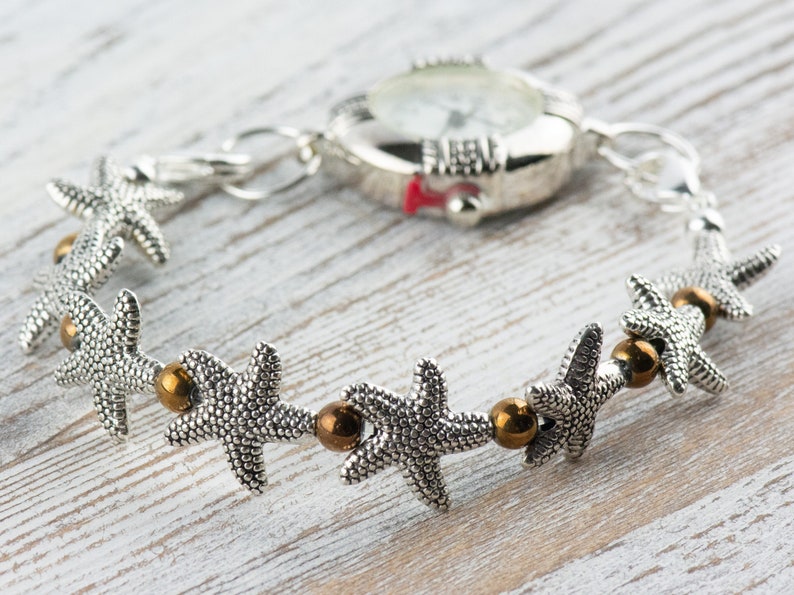 K&ouml;nnte beinhalten: Eine silberne Uhr mit wei&szlig;em Zifferblatt und einem silbernen Armband, das mit Seestern-Anh&auml;ngern und bronzenen Perlen verziert ist.