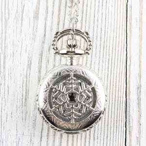 May include: A silver pocket watch with a snowflake design on the face. The watch is on a chain and hanging against a white wooden background.