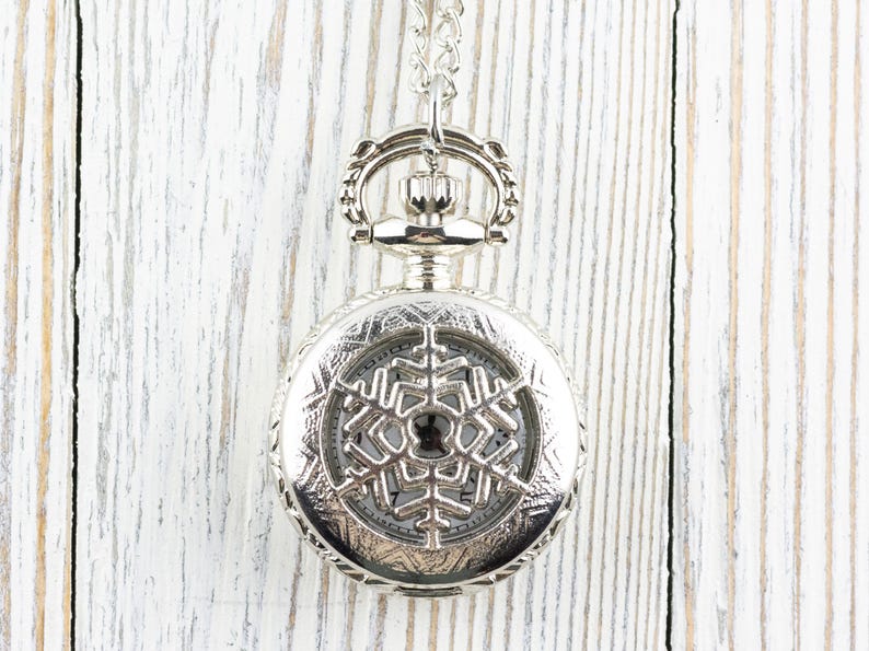 May include: A silver pocket watch with a snowflake design on the face. The watch is on a chain and hanging against a white wooden background.