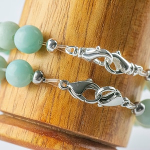 May include: Two strands of a light green beaded bracelet with silver clasps. The bracelet is displayed on a wooden surface.