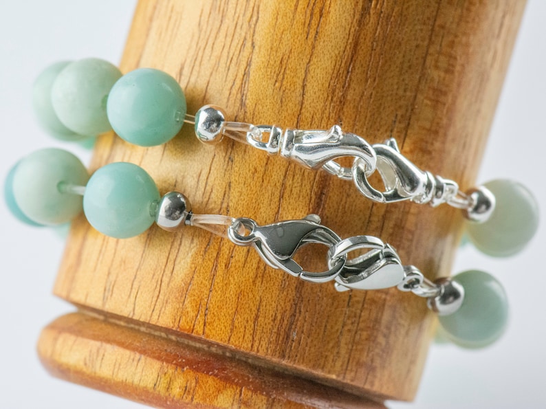 May include: Two strands of a light green beaded bracelet with silver clasps. The bracelet is displayed on a wooden surface.