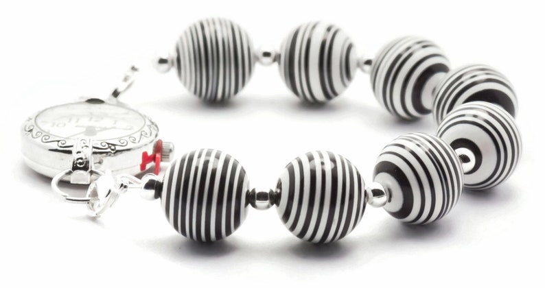 K&ouml;nnte beinhalten: Ein Armband aus schwarzen und wei&szlig;en Streifen mit einem silbernen Zifferblatt. Das Armband besteht aus runden Perlen mit einem schwarzen und wei&szlig;en Streifenmuster.