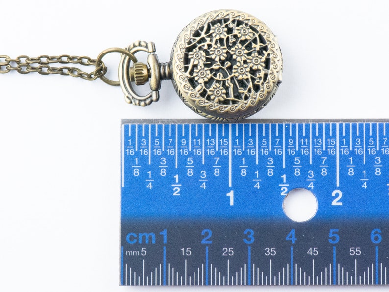 K&ouml;nnte beinhalten: Antike bronzefarbene Taschenuhr mit einem verzierten Blumendesign auf dem Deckel. Die Uhr ist an einer passenden Kette befestigt. Die Uhr hat einen Durchmesser von ca. 5 cm, wie auf dem Lineal gezeigt.