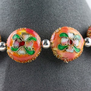 May include: A close-up of a bracelet with round, orange beads decorated with green and white floral designs. The beads are separated by silver spacer beads.
