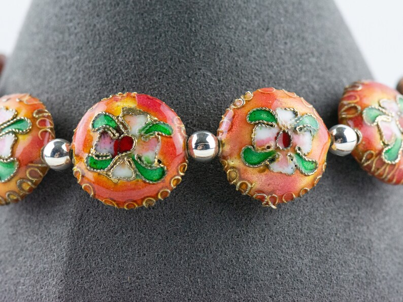 May include: A close-up of a bracelet with round, orange beads decorated with green and white floral designs. The beads are separated by silver spacer beads.