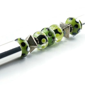 May include: A silver pen with a green and black patterned glass bead, a silver spacer bead, and a clear glass bead with green and white polka dots.