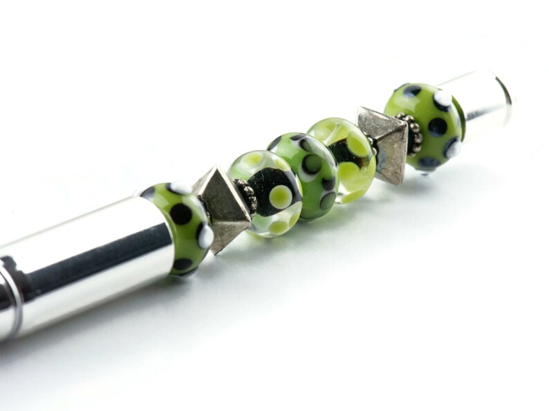 May include: A silver pen with a green and black patterned glass bead, a silver spacer bead, and a clear glass bead with green and white polka dots.