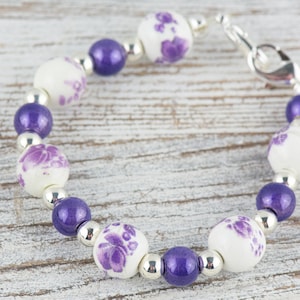 May include: A purple and white beaded bracelet with a silver clasp. The beads are round and have a floral design.