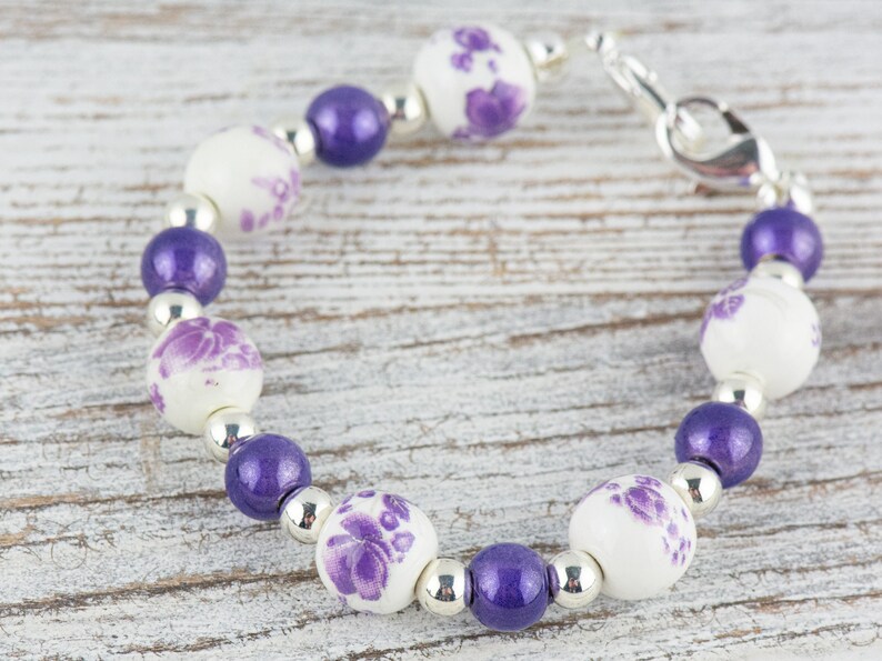 May include: A purple and white beaded bracelet with a silver clasp. The beads are round and have a floral design.