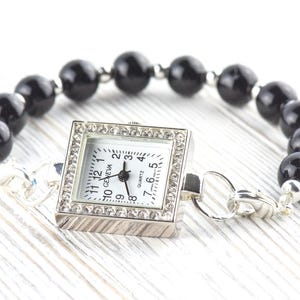 May include: A silver-toned quartz watch with a square face, surrounded by small crystals, is attached to a bracelet of large, black, round beads. The watch face has black numerals and hands. The bracelet is on a white wooden surface.