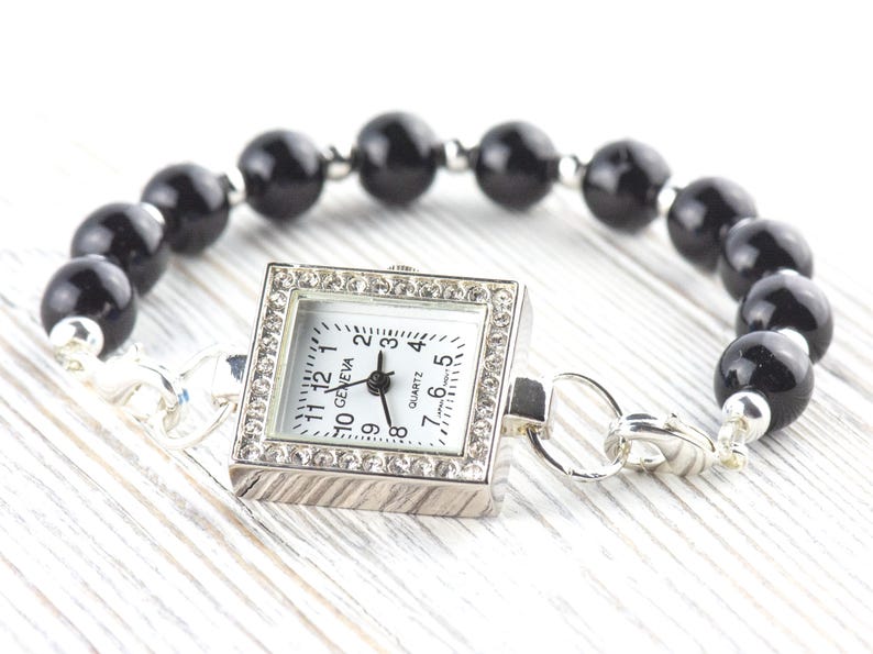 May include: A silver-toned quartz watch with a square face, surrounded by small crystals, is attached to a bracelet of large, black, round beads. The watch face has black numerals and hands. The bracelet is on a white wooden surface.