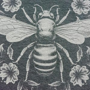May include: A detailed illustration of a bee with outstretched wings, surrounded by floral and leaf elements. The design is in a light gray color, set against a dark gray background, creating a striking contrast. The artwork has a vintage, botanical theme.