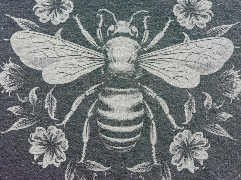 May include: A detailed illustration of a bee with outstretched wings, surrounded by floral and leaf elements. The design is in a light gray color, set against a dark gray background, creating a striking contrast. The artwork has a vintage, botanical theme.