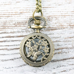 May include: A gold-toned pocket watch necklace with a delicate floral design and a butterfly in the center. The watch face is visible through the intricate design.