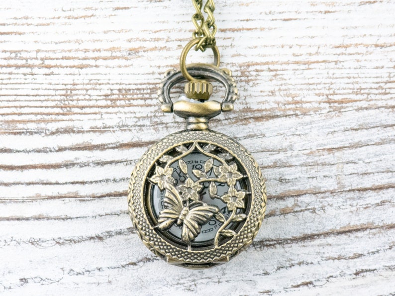 May include: A gold-toned pocket watch necklace with a delicate floral design and a butterfly in the center. The watch face is visible through the intricate design.