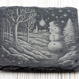 K&ouml;nnte beinhalten: Ein schwarzer Schieferuntersetzer mit einem wei&szlig;en, ge&auml;tzten Design eines Schneemanns in einer Winterlandschaft. Der Schneemann tr&auml;gt einen Hut und einen Schal und steht in einem verschneiten Wald mit B&auml;umen und einem kleinen Geb&auml;ude im Hintergrund.