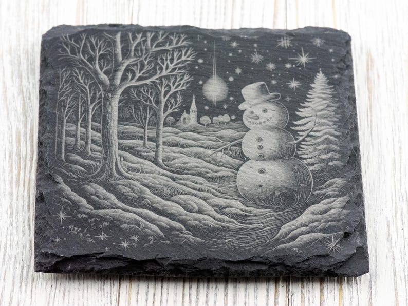 K&ouml;nnte beinhalten: Ein schwarzer Schieferuntersetzer mit einem wei&szlig;en, ge&auml;tzten Design eines Schneemanns in einer Winterlandschaft. Der Schneemann tr&auml;gt einen Hut und einen Schal und steht in einem verschneiten Wald mit B&auml;umen und einem kleinen Geb&auml;ude im Hintergrund.