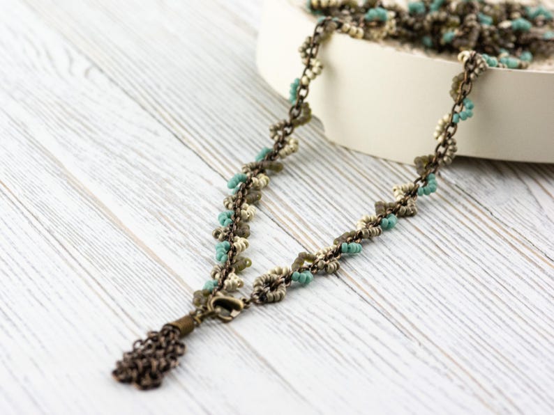 May include: A delicate necklace with a tassel made of small, turquoise, beige and brown beads. The necklace is made of metal and has a chain-like design.