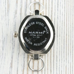 May include: A silver pocket watch with a black face. The watch has a stainless steel back and is water resistant. The face of the watch has the brand name "NARMI" and the text "JAPAN, MOVT L-39". The watch has two silver rings for attachment.