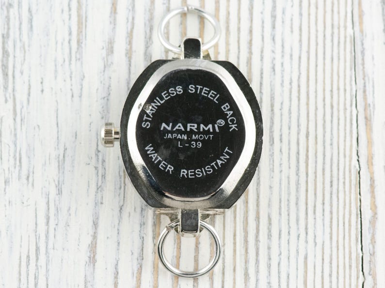 May include: A silver pocket watch with a black face. The watch has a stainless steel back and is water resistant. The face of the watch has the brand name "NARMI" and the text "JAPAN, MOVT L-39". The watch has two silver rings for attachment.