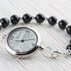 Puede incluir: Un reloj de pulsera plateado con una esfera blanca redonda que presenta contornos de mariposas negras y el texto Geneva. El reloj tiene una correa hecha de cuentas esf&eacute;ricas negras pulidas separadas por peque&ntilde;os espaciadores met&aacute;licos plateados.