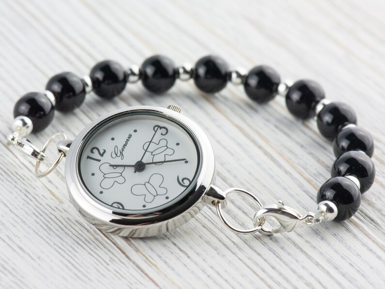 Puede incluir: Un reloj de pulsera plateado con una esfera blanca redonda que presenta contornos de mariposas negras y el texto Geneva. El reloj tiene una correa hecha de cuentas esf&eacute;ricas negras pulidas separadas por peque&ntilde;os espaciadores met&aacute;licos plateados.