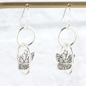 May include: Silver dangle earrings featuring a butterfly design. Each earring has a circular top and a butterfly charm suspended within an oval-shaped frame. The butterflies have detailed wing patterns.