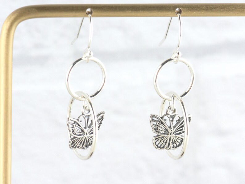 May include: Silver dangle earrings featuring a butterfly design. Each earring has a circular top and a butterfly charm suspended within an oval-shaped frame. The butterflies have detailed wing patterns.