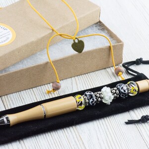 May include: A wooden pen with a silver tip and a beaded charm bracelet attached. The pen is in a black velvet pouch and the charm bracelet is in a brown cardboard box with a yellow ribbon and a heart-shaped charm.