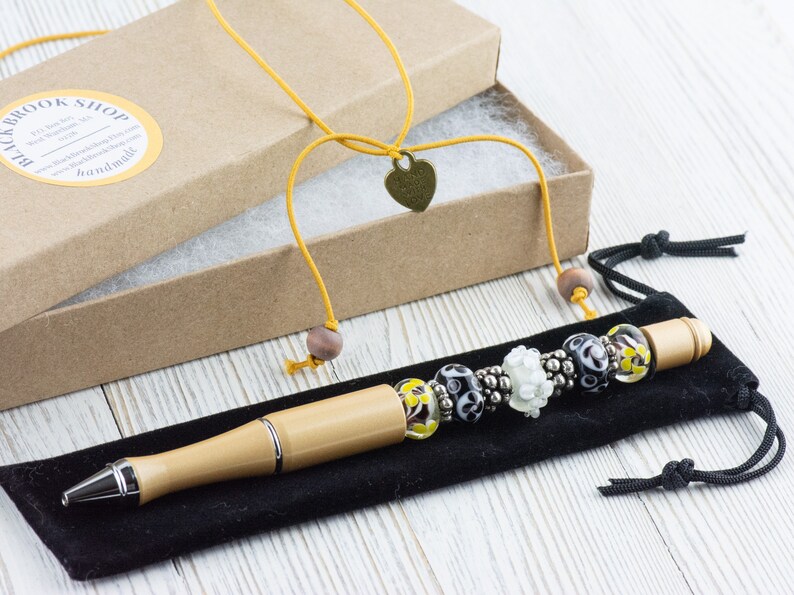 May include: A wooden pen with a silver tip and a beaded charm bracelet attached. The pen is in a black velvet pouch and the charm bracelet is in a brown cardboard box with a yellow ribbon and a heart-shaped charm.