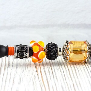 May include: A black pen with decorative beads. The beads include a black and silver bead with black stones, an orange and white bead, a black bead, and a yellow rectangular bead with silver filigree.