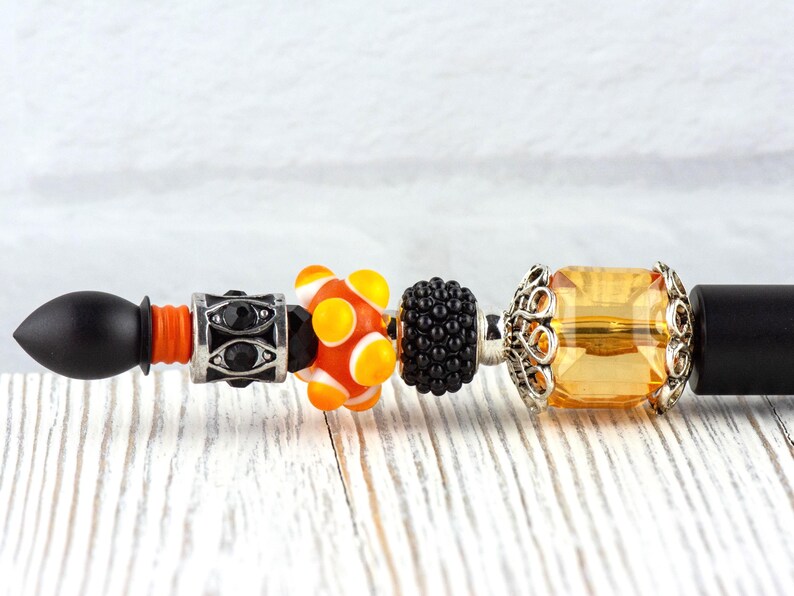 May include: A black pen with decorative beads. The beads include a black and silver bead with black stones, an orange and white bead, a black bead, and a yellow rectangular bead with silver filigree.