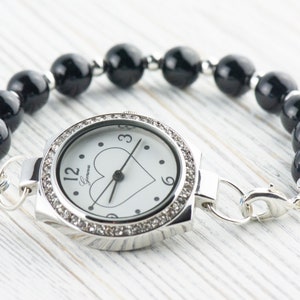 May include: A silver watch with a white face and black heart design. The watch is attached to a black beaded bracelet.