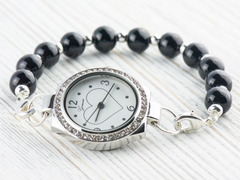 May include: A silver watch with a white face and black heart design. The watch is attached to a black beaded bracelet.