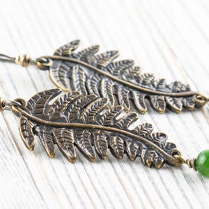 K&ouml;nnte beinhalten: Ein Paar bronzefarbene Ohrringe in Form von Farnbl&auml;ttern mit gr&uuml;nen Jade-Steinen. Die Ohrringe sind im H&auml;nge-Stil und haben einen rustikalen, nat&uuml;rlichen Look.