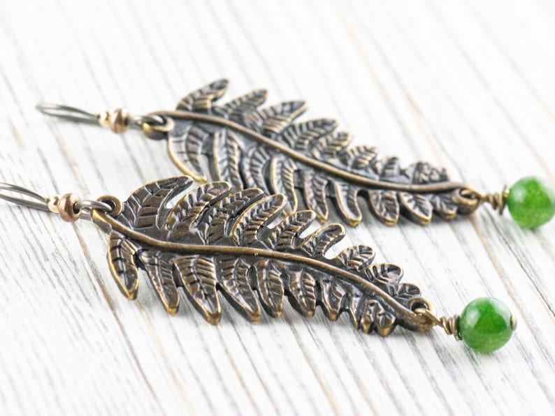 K&ouml;nnte beinhalten: Ein Paar bronzefarbene Ohrringe in Form von Farnbl&auml;ttern mit gr&uuml;nen Jade-Steinen. Die Ohrringe sind im H&auml;nge-Stil und haben einen rustikalen, nat&uuml;rlichen Look.