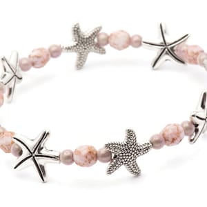 K&ouml;nnte beinhalten: Ein Stretch-Armband mit silbernen Seestern-Anh&auml;ngern und rosa und malvenfarbenen Perlen. Das Armband-Design wechselt zwischen Seesternen und runden Perlen und schafft ein maritimes Thema. Die Perlen haben ein gesprenkeltes Aussehen.