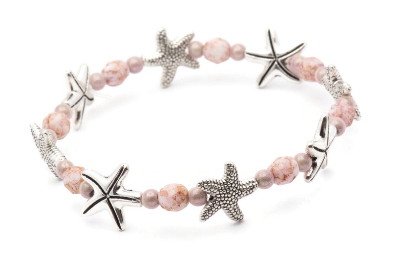 K&ouml;nnte beinhalten: Ein Stretch-Armband mit silbernen Seestern-Anh&auml;ngern und rosa und malvenfarbenen Perlen. Das Armband-Design wechselt zwischen Seesternen und runden Perlen und schafft ein maritimes Thema. Die Perlen haben ein gesprenkeltes Aussehen.
