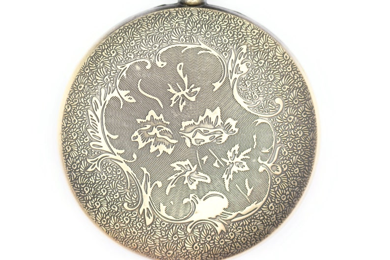 K&ouml;nnte beinhalten: Ein runder Anh&auml;nger aus Messing mit einem filigranen Blumenmuster. Das Design zeigt ein sich wiederholendes Muster aus Bl&auml;ttern und Spiralen.
