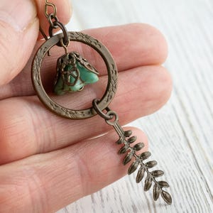May include: A bronze-coloured earring with a teal stone pendant and a fern-shaped charm. The earring has a circular design with detailed patterns. The earring is held in a person's hand.